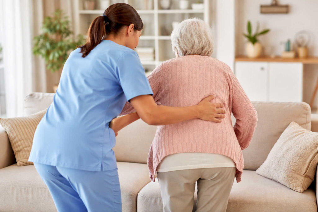 Badante che aiuta una signora anziana ad alzarsi dal divano in casa, assistenza domiciliare ripresa di spalle