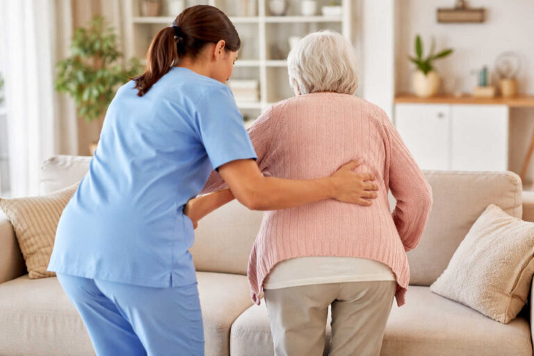 Badante che aiuta una signora anziana ad alzarsi dal divano in casa, assistenza domiciliare ripresa di spalle